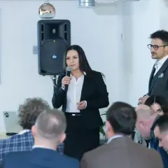 Kobieta w garniturze przemawia do grupy ludzi, obok mężczyzna w okularach.