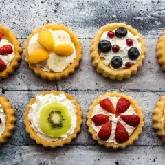 Osiem kolorowych tartaletek z owocami: truskawkami, borówkami, kiwi, bananami i morelami na kremowym nadzieniu.