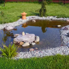 Ogród z oczkiem wodnym, kamieniami, roślinnością i ceramiczną latarenką.