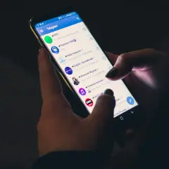 Dłonie trzymają smartfon z otwartą aplikacją Telegram. Czy telegram jest bezpieczny? Aplikacja wyświetla listę czatów i kanałów.