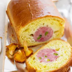 Un saucisson brioché doré, coupé en tranches, révèle sa garniture rosée. Des girolles et un couteau complètent cette gourmandise.