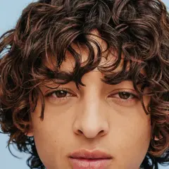 Gros plan sur le visage d'un homme aux cheveux bruns ondulés, type cheveux 2b homme, sur fond bleu clair.