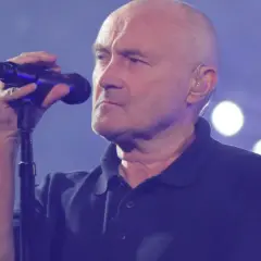Phil Collins, wokalista zespołu Genesis, śpiewa do mikrofonu na tle niebieskiej sceny z białymi światłami.