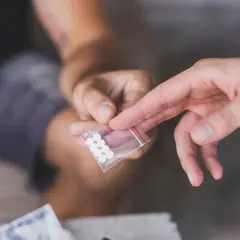 Dwie dłonie, jedna podaje, druga odbiera małą torebkę z białymi tabletkami, być może mdma.