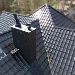 Nowoczesny, czarny komin stalowy na błyszczącym, ciemnym dachu. W tle drzewa.
