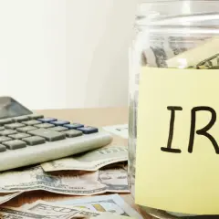 A glass jar labeled "IRA" sits on a table with US dollar bills and a calculator, symbolizing financial planning and the potential for an inherited IRA.