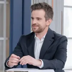 Łukasz Jedliński, dziennikarz TVN, w garniturze siedzi przy stole z dokumentami i szklanką wody.