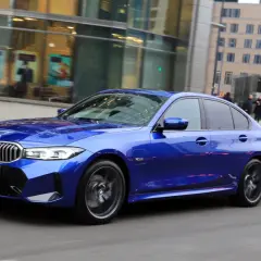 Błękitne BMW 330e mija nowoczesne budynki i wejście do metra.