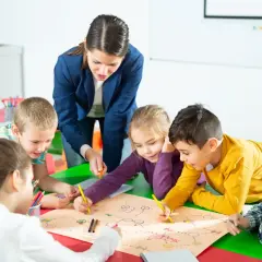 Nauczycielka z przedszkola publicznego pomaga dzieciom w rysowaniu mapy. Warto wiedzieć, ile kosztuje godzina w przedszkolu publicznym.