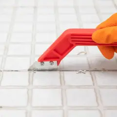 Mano con guanto arancione usa uno strumento rosso per rimuovere lo stucco tra le piastrelle, un passo fondamentale per capire come togliere le piastrelle dal muro.