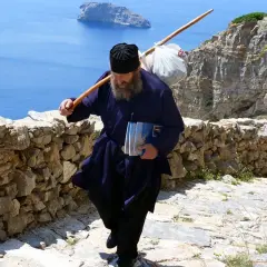En munk vandrar uppför en stenstig på Amorgos, Grekland, med en stav över axeln och böcker i handen.
