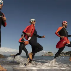 Ludzie w piankach do pływania biegają po plaży z pomarańczowymi bojami.