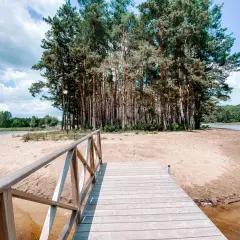 Drewniany pomost prowadzi do piaszczystej plaży otoczonej lasem sosen.