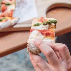 Eine Hand hält ein Stück Pinsa mit Tomaten, Mozzarella und Basilikumpesto. Was ist Pinsa? Eine römische Spezialität, die sich von Pizza unterscheidet.