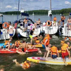 Ludzie w kamizelkach ratunkowych i strojach kąpielowych pływają i bawią się na deskach SUP przy żaglówkach. Wspaniały weekend czerwcowy na wodzie.