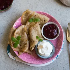 Glutenfria pannkakor med sylt och grädde på en rosa tallrik. En gaffel och kniv ligger bredvid.