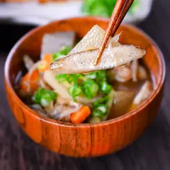 Chopsticks lift a piece of daikon from a steaming bowl of Japanese vegetable soup, filled with carrots, mushrooms, and green onions.