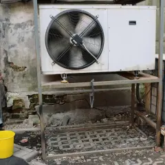 Dwa urządzenia klimatyzacyjne stoją na zewnątrz, jedno większe z wentylatorem, drugie mniejsze. Obok żółte wiadro.
