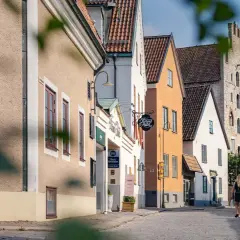 Ett par promenerar hand i hand på en kullerstensgata i Visby, förbi charmiga hus och skyltar som indikerar B&B Gotland och Strand Hotel.