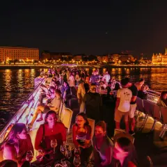 Nocny rejs po Dunaju w Budapeszcie. Ludzie bawią się na statku, w tle oświetlony Parlament.