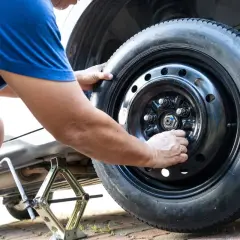 Un homme ajuste les écrous d'une roue de secours sur une voiture. Le cric est visible sous le véhicule.