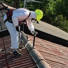 Pracownik w białym kombinezonie i żółtym kasku usuwa stare pokrycie dachowe, prawdopodobnie eternit, z czerwonego dachu.