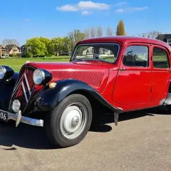 Klasyczny, czerwony Citroen Traction Avant, jeden z kultowych modele stare, zaparkowany na tle zielonej łąki i domów.