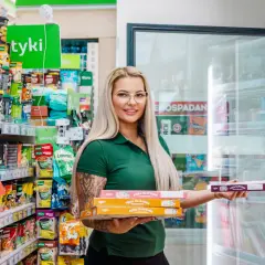 Kobieta w zielonej koszulce trzyma stos pudełek z pizzą na wynos w sklepie osiedlowym.