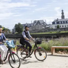 Couple à vélo profitant d'une balade ensoleillée le long de la rivière, avec les bâtiments historiques de Tréguier Lannion en arrière-plan.