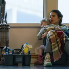 Madre abbraccia figlia, circondata da attrezzi per le pulizie e un aspirapolvere. Le recensioni per il suo lavoro di maid sono positive, ma la vita è dura.