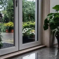 Przez nieszczelne drzwi wejściowe widać deszczowy ogród z kwitnącymi kwiatami i zielenią. Wewnątrz stoi duża roślina doniczkowa.