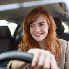 Donna sorridente al volante, con capelli rossi e lentiggini. Pensa alla sua assicurazione giornaliera auto per un viaggio sereno.