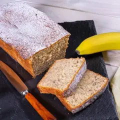 Saftiger Bananenkuchen, bestäubt mit Puderzucker. Daneben liegen zwei Scheiben und eine reife Banane – der beste Bananenkuchen der Welt!
