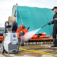 Hombre con traje de trabajo y botas limpiando maquinaria agrícola con hidrolimpiadora. Aprende como usar hidrolimpiadora para dejar todo impecable.