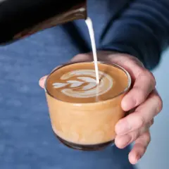 En hand häller mjölk i en kopp för att skapa ett vackert mönster på en flat white.