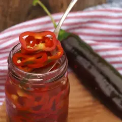 Burk med picklad chili, en sked med chiliringar och en hel mörk chili i bakgrunden.