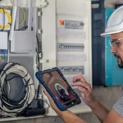 Technik w kasku i okularach ochronnych wykonuje przegląd instalacji elektrycznej domu, korzystając z tabletu z aplikacją diagnostyczną.