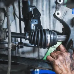 Mechanik z brudnymi rękami wyciąga półoś ze skrzyni biegów, widoczny przegub i osłonę gumową.