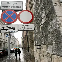 Znaki drogowe informują o zakazie zatrzymywania się i postoju na drodze wewnętrznej. W tle widać ulicę z samochodami i przechodniów.
