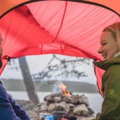 Ett par skrattar inne i ett orange tält. Utanför syns en sjö, träd och en lägereld. Perfekt för att campa i skogen.