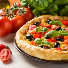 Domowa pizza z pomidorami, oliwkami i kukurydzą, gotowa do pieca. Obok świeże pomidory i bazylia.