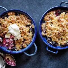 Två blåa ugnsformar med glutenfri smulpaj fylld med bär och toppad med grädde. Serveras med antika skedar.