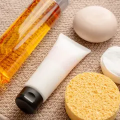 Płyn do kąpieli z **sodium lauryl sulfate**, krem, mydło, waciki i gąbka do twarzy na ręczniku.