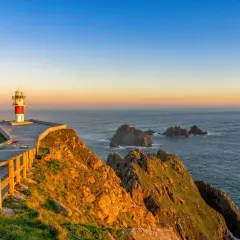 Faro en el cabo Ortegal al atardecer, con el mar azul y rocas emergiendo de las olas.