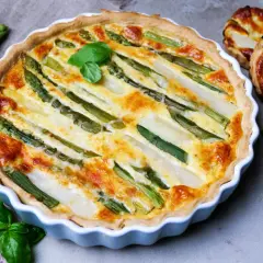 Herzhafte Quiche mit Spargel, frisch gebacken und mit Basilikum garniert. Daneben liegen frischer Spargel und kleine Spargel-Törtchen.