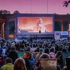 Stor publik njuter av sommarbio Stockholm under en lila kvällshimmel. En film visas på en stor duk framför tält och träd.