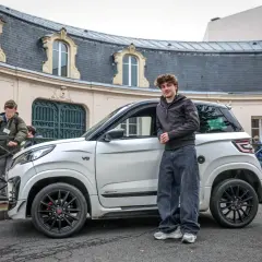 Un jeune homme pose à côté d'une voiturette électrique blanche. Le **rayon de braquage** de ce véhicule compact est idéal pour les rues étroites.
