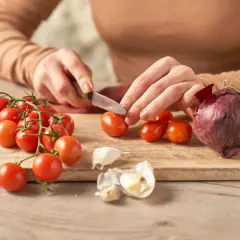 Hände schneiden Tomaten für selbstgemachten Ketchup. Zwiebel und Knoblauch liegen bereit.