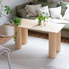 Ein heller Holztisch mit säulenartigen Beinen, perfekt für ein DIY-Projekt. Daneben ein grünes Sofa, Pflanzen und ein Körbchen.