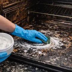 Mano con guanto blu pulisce un forno incrostato con una spugna e una pasta bianca.